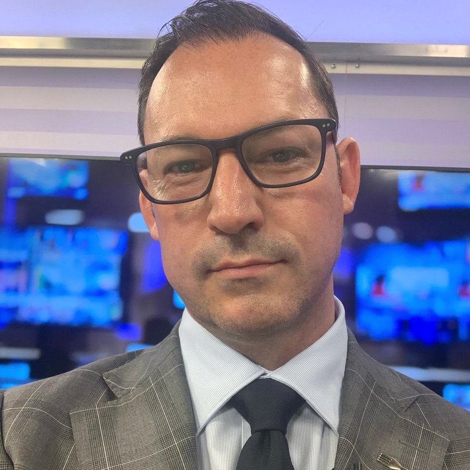 Wes Lambert wearing glasses and a grey suit, with blue screens behind him